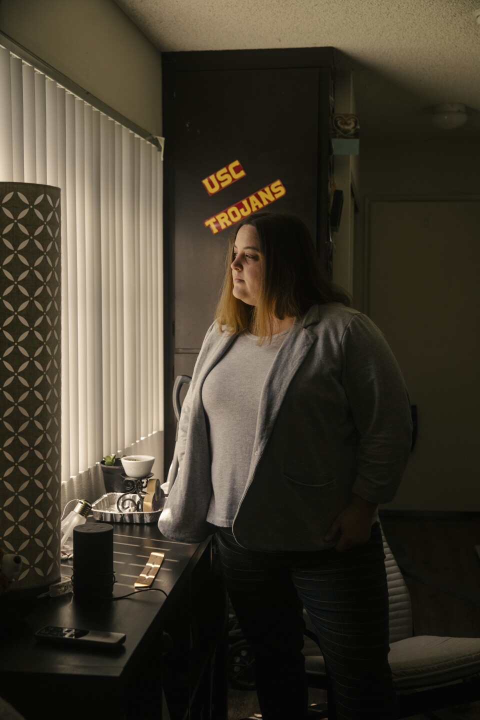 A woman with mid-length brown hair wearing a gray blazer and black pants stands in a kitchen in front of the window with the blinds drawn. On the cupboard behind her, there's a sticker that says "USC Trojans."