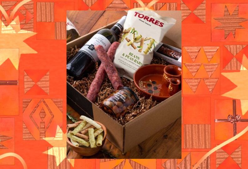 A gift box containing a bottle of red wine, two types of dried cured meats, a jar of olives and an olive bowl, and a bag of crunchy snacks — all the making of a tapas party. The photo is set against a decorative orange-and-gold background, accented with stars, holiday gift boxes and strands of ribbon.