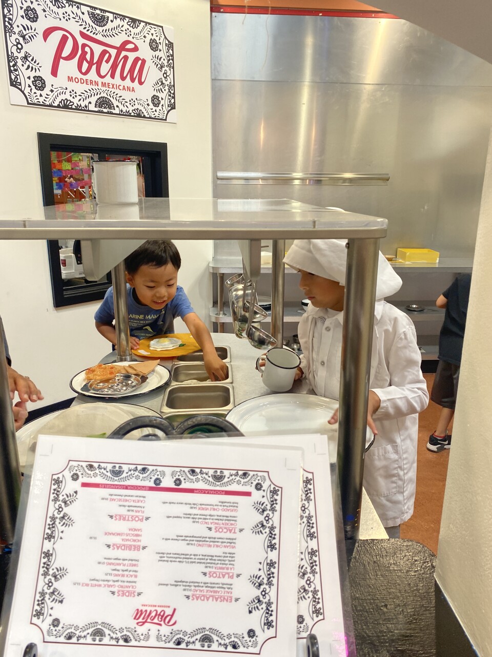 A boy in a blue shirt and another boy in a white chef's hat and white apron lean next to a warming table, pretending to plate up food. 