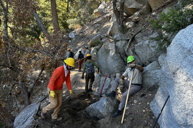 Multiple younger and middle-aged men on a dirt trail with large boulders wearing hardhats and holding McLeod tools do work on a trail. 