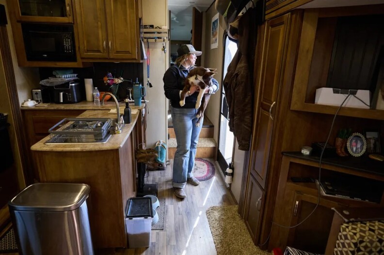 A woman wearing a baseball cap, jeans and dark sweater holds a small dog in her arms as she looks out the door of an RV.