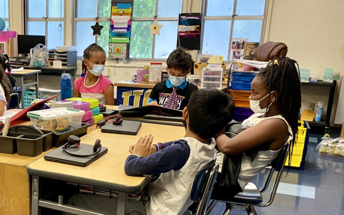 Four elementary school students, all wearing face masks, sit around a table in a classroom crowded with desks, bins of supplies and wall art.
