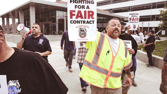 A man in a neon vest is walking and holding a sign that reads "For a Fair Contract Teamsters Local 952". Crowds of people behind him are also walking and holding signs.