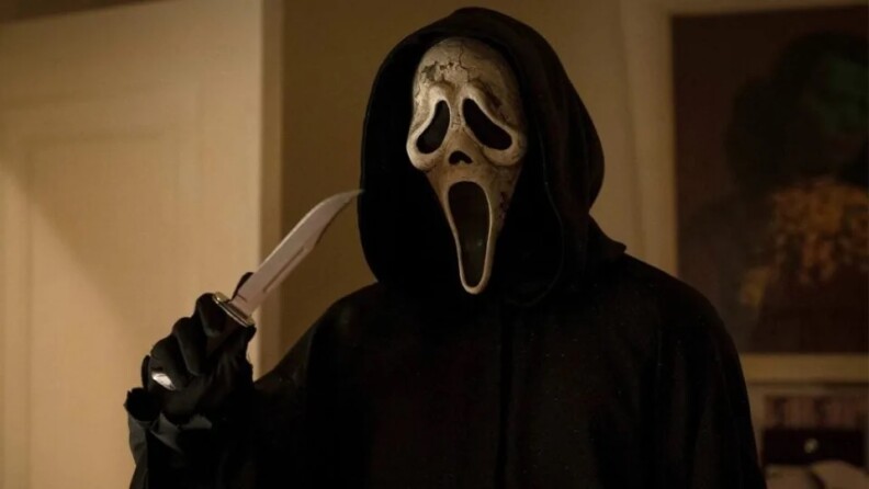 A person in a black cloak and white mask stands in a living room. The black cloak covers the person's entire body with a hood pulled up over the person's head and black gloves covering the person's hands. The white mask has a fixed, cartoonish expression that resembles a generic ghost face. The mouth, nose, and eyes are indicated with blacked out dents. The mouth shape is an exaggerated skinny oval that signifies a scream. The nose is indicated by a small black nostril hole similar to that of a skull. And the eye dents are large oblong shapes that give the expression of a furrowed sorrowful brow. The mask has dirt splotches on it with some of white paint wearing off. The person holds a large hunting knife in their right hand about shoulder height with the tip pointing slightly to the mask as if gesturing towards themselves. 