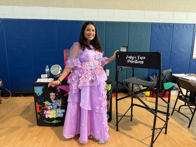 A brown skinned woman in a purple tulle dress next to a chair that says "Zuly's face painting" in a gym. Her art supplies are in the background.