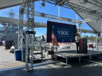 A female presenting person in slacks and a white shirt speaks at a podium in front of a blue screen with the letters VBOC. An American flag is visible on the stage. Scaffolding as well as a battle ship is visible in the background. 