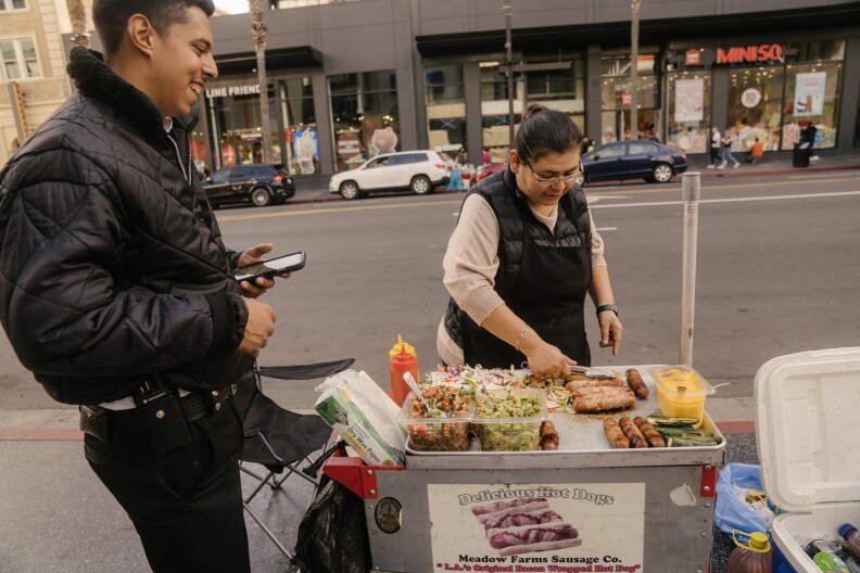 A man in a black puffy jacket, at left, stands next to a cart where a woman in a black vest cooks hot dogs, with a wide street behind them.