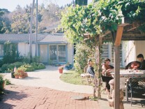 A family sits outside under an awning covered in green vines. It's a sunny day. A house and palm trees are in the background.