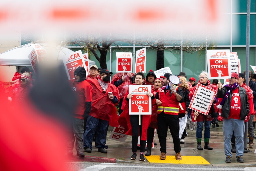 A group of people wearing red stand on a sidewalk holding red and white signs that read "CFA/ ON STRIKE!" with a symbol of a hand in a fist holding a pencil.