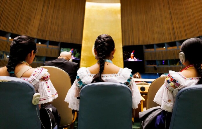 Three women are pictured from behind while sitting in blue chairs in a large, circular room. They each wear their hair in a single braid and are wearing white dresses with colorful embroidery. 