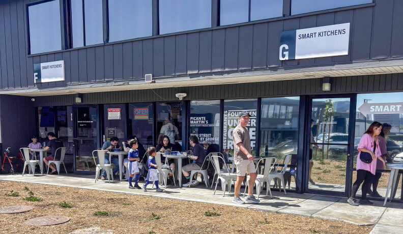 Exterior of a business park building with "Smart Kitchens" signage above glass doors, with customers seated at outdoor tables and pedestrians walking past on the sidewalk.