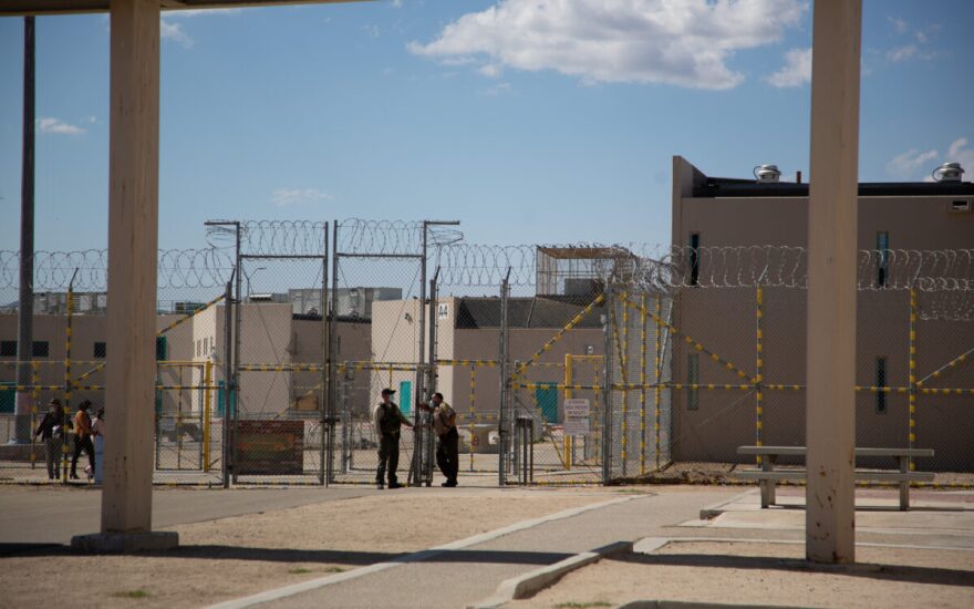 A drab facility surrounded by tall chain-link fences that are topped with spiraling barb wire. A couple of guards stand in front of a gate and some other people are walking behind the fenceline.