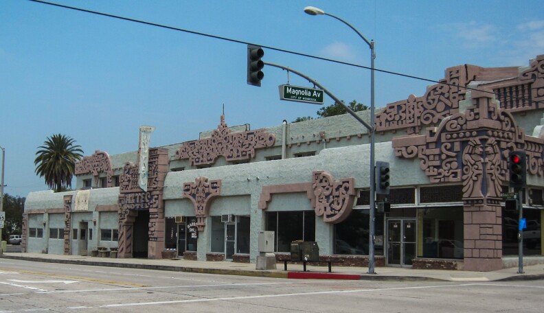 A wide shot of the street corner for the Aztec Hotel, which is a complex that takes up the entire corner. The streetlight for Magnolia Avenue is shown. The shop spaces in view appear empty.