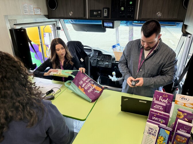 A  man and a woman with medium light skin tone sit behind a green counter. There are bookshelves lining either side and a clear windshield behind them. The woman places a book in a teal and purple bag. 