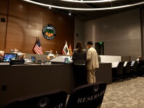 An older man wearing a beige vest and black hat stands next to a woman in a gray blazer as they stand behind a microphone and speak towards people sitting behind a gray dais. 