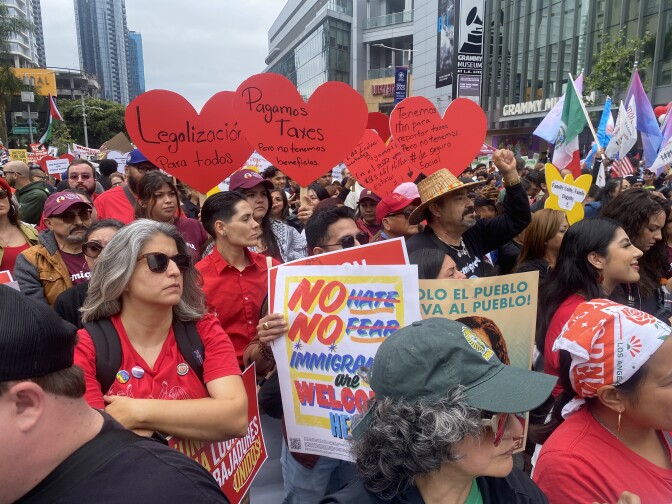 A crowd of people at a protest are carrying red heart signs and colorful flags on a city street.