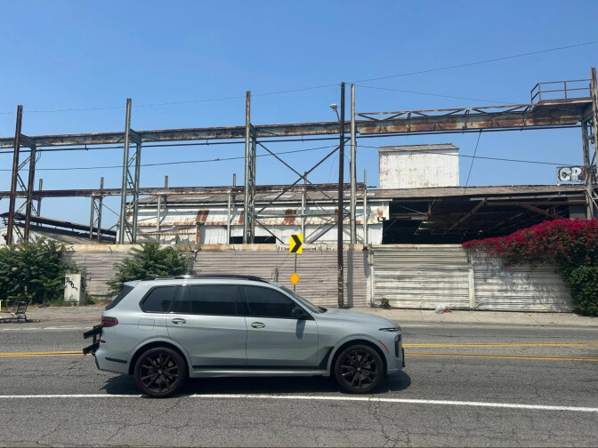A car passes by a facility with rusted walls and walkways.