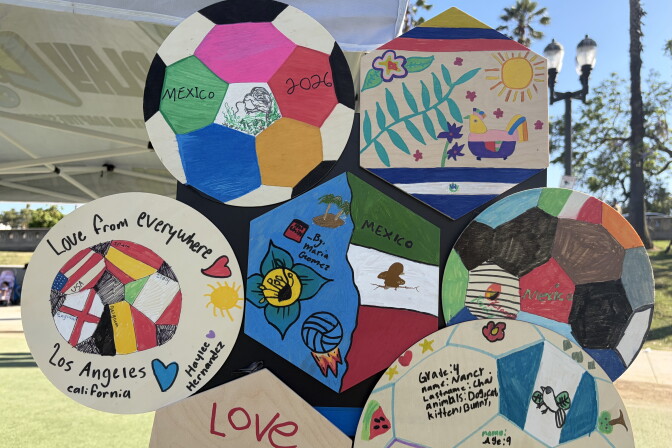 A handful of wooden circles and hexagons, decorated in vibrant colors by children, are affixed to a temporary display board at a park. Palm trees and street lamps cans be seen in the background. 