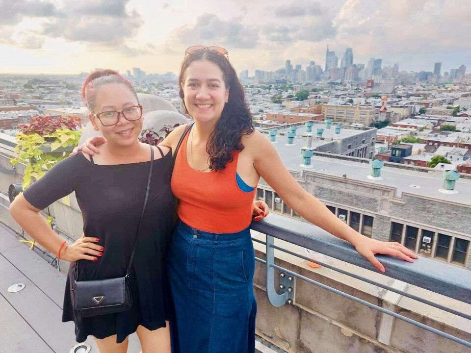 Two women of Asian descent pose on a rooftop with a cityscape in the background.