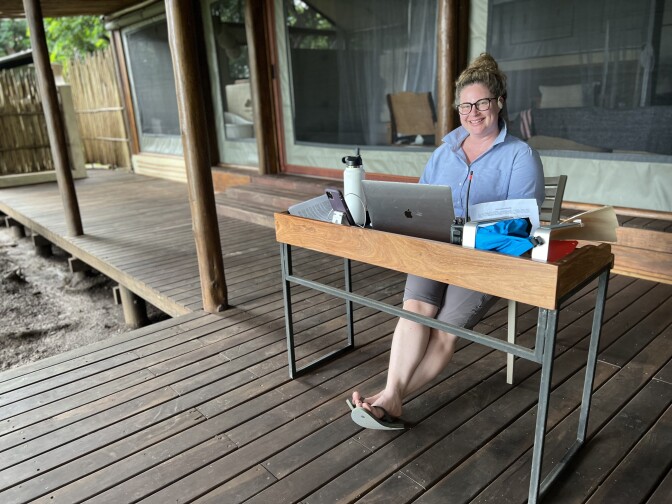 A white woman wearing glasses, a blue shirt and a tousled bun, sits at a desk outdoors on a deck.