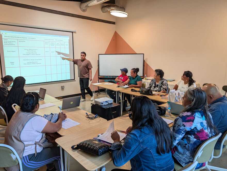 Ten people sit in a classroom. They look at a person standing, pointing to an image on a screen.