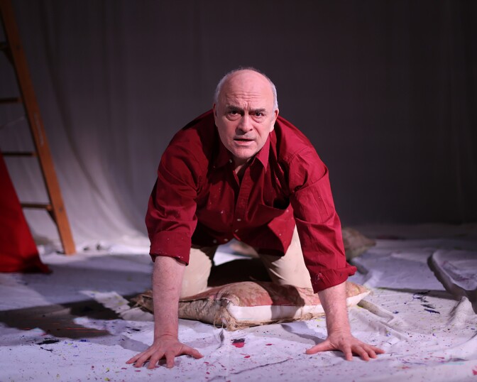 A light-skinned bald man in a red shirt kneels on all fours on a pillow on top of an artist's canvas. 