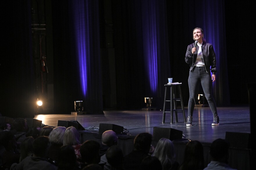 A stand up comedian stands on stage, sporting a black jacket, pants, shoes, and white t-shirt.