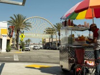 A street vendor stands under a rainbow umbrella peeling fruit in front of a cart. In the distance is an arch with a sign that reads "Whittier Blvd. East Los Angeles."