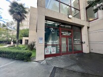 We're looking at the entrance to the downtown jail public lobby and inmate reception center. A concrete beige building faces us, with glass windows framed with red. The door has white signs like "put on your mask". The building is part of a large complex -- we can see other beige buildings beyond the reception center. A palm tree is to the right of the building, and there are green grass and shrubs. 