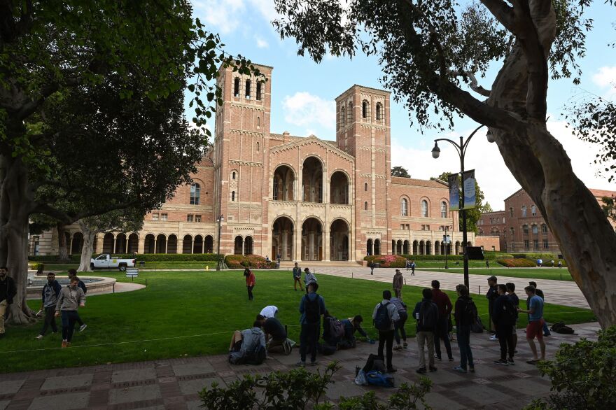 A red-brick building has two towers on either side of an arched entry. Students sit and stand on campus walkways in the foreground.