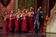 A group of singers in two rows on a stage, with arms crossed in front of them and wearing red robes and hats, both with gold accents. To their left is singer-songwriter Josh Groban in a medeival style outfit with partial knight armor, with his arms outstretched and mouth open in song.