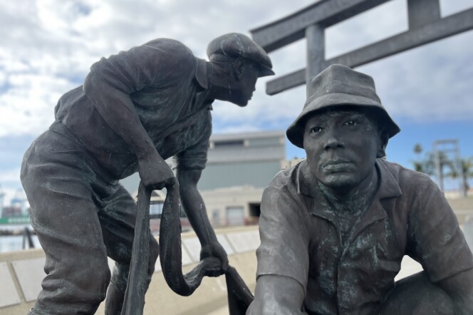 A bronze statue of two workers. One worker is standing and holding a rope, while the other is kneeling. In the background, there are industrial buildings.