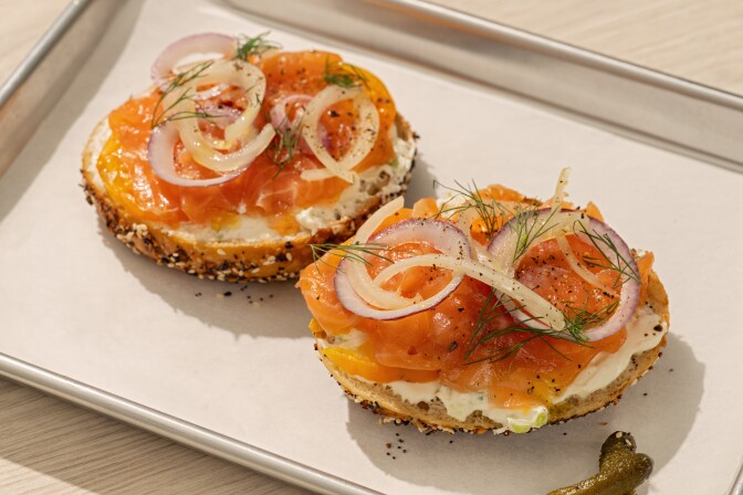 Open-face everything bagel halves topped with cream cheese, yellow tomato slices, smoked salmon, pickled red onions, fresh dill, and black pepper on a metal tray. 