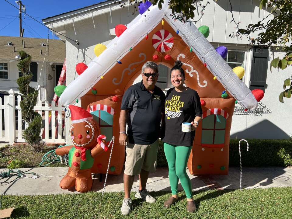 A man wearing a black t-shirt and khaki shorts and a woman wearing green leggings and a black t-shirt stand in front of an inflatable gingerbread house. 