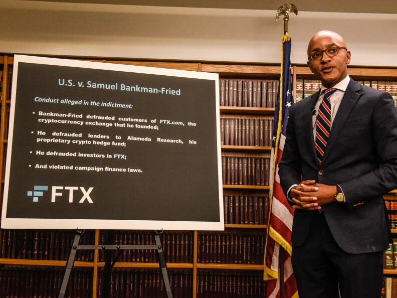 A bald man with light-brown skin tone, wearing glasses and dressed in a suit, stands off to right and next to a sign on a stand that lists a variety of bullet points. At the top of the sign reads "U.S. v. Samuel Bankman-Fried."