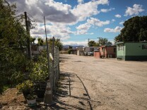 An image of a deserted dirt road. A chain link fence borders the road on the left, and several mobile homes are on the right. One is green, one is red and one is white. It is day time and the sky is white with clouds. 