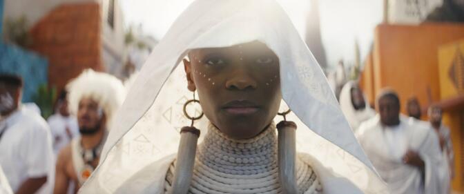 Letitia Wright stands center frame, only her shoulders up visible. She wears all white, her head hooded by a shear white fabric that falls past her shoulders. Her neck is wrapped in white beaded necklaces starting at her chest and raveling all the way up to her chin. She also wears large earrings that look like saber teeth on each ear and they too fall past her shoulders. She stares straight ahead, her face sullen. White paint is dotted into fine lines across her cheekbones and up her nose. Behind her walks a crowd of other people, also in white as if in a procession. 