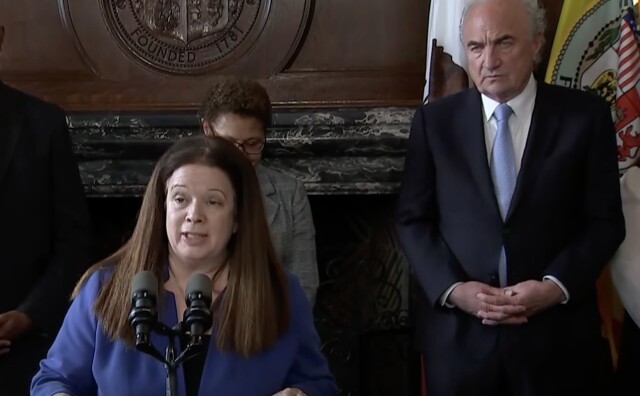 City Attorney Hydee Feldstein Soto, a woman with light skin tone, straight brown hair, wearing a blue blazer stands at a podium and speaks into a microphone. Behind her is Mark Adams, a man with light skin tone, white hair, wearing a navy blue blazer, white shirt, and light blue tie. He stands in front of two flags. 