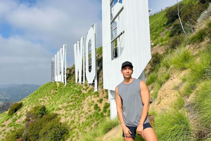 A light-skinned Latino man in a baseball hat, tank top and shorts stands just inches away from The Hollywood Sign