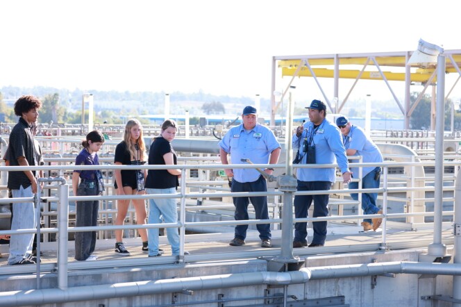 A group of high school students hear from adult water professionals in light blue attire at a water treatment facility outside on a sunny day. 