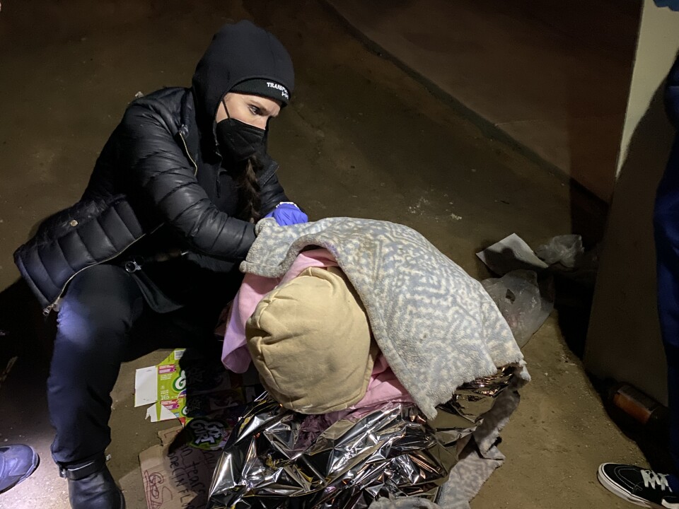 A woman in a black jacket, beenie and wearing a face mask kneels next to a person with a blanket over their back and their hoodie-covered head bowed. 