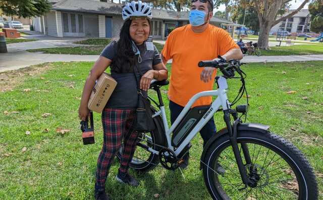 A Latino couple pose while holding their new e-bike in the middle of a park.