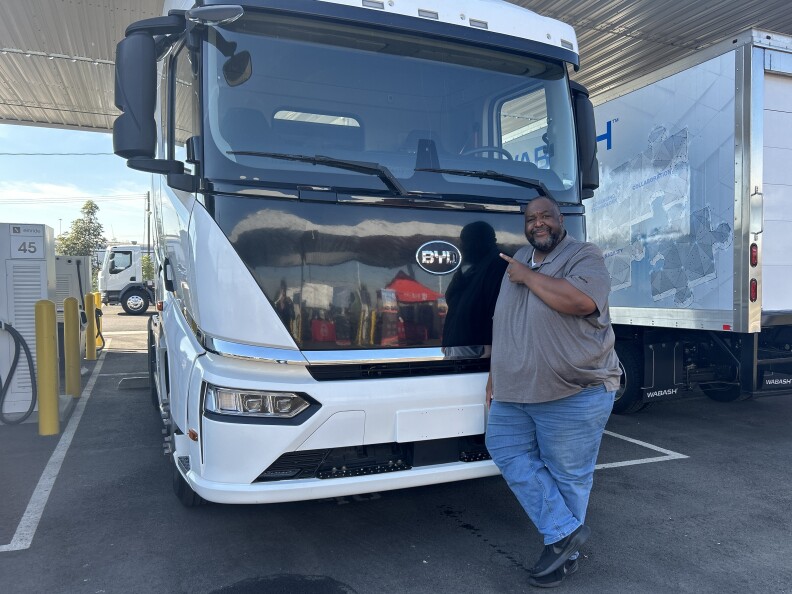 A Black man with a bald head and short black/grey mustache and bears stands in front of a large black and white BYD flat-faced electric truck. He wears a grey collared t-shirt and jeans and points to the BYD brand in the center of the truck and smiles