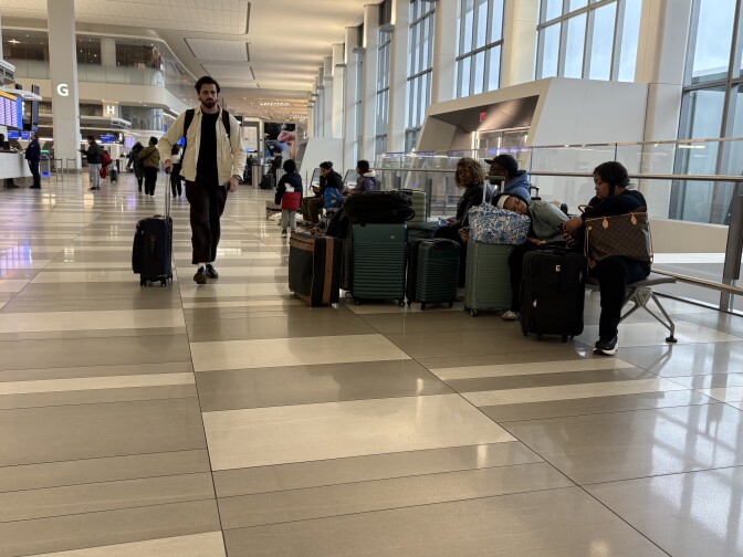 People sitting at an airport with luggage in front of them. A man pushing a luggage walks past the seated people.