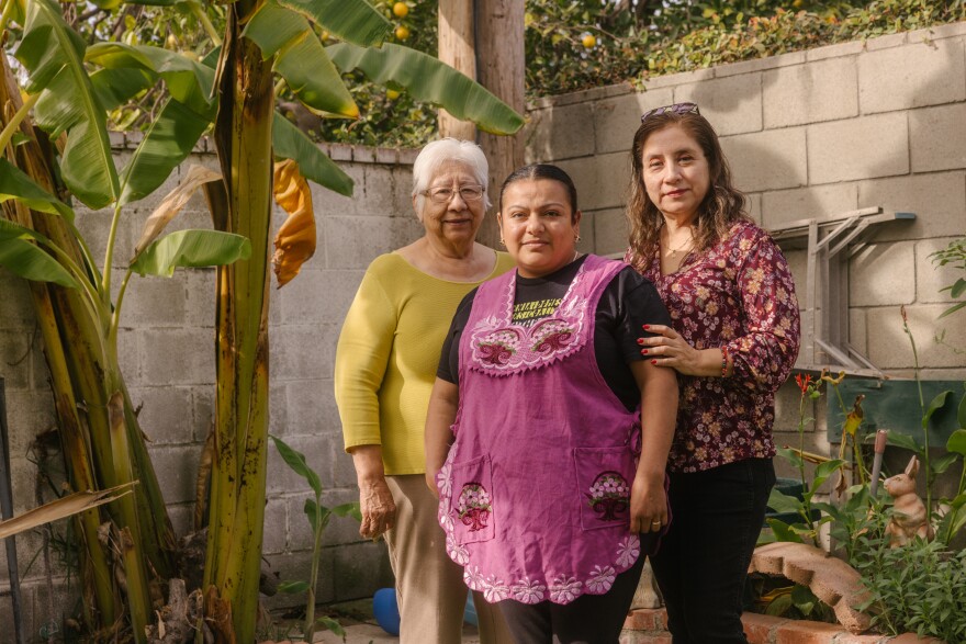 An older woman with medium-light skin tone and short white hari wearing a yellow long sleeve shirt, beige pants, and glasses stands next to a woman with medium skin tone with black hair tied in a bun wearing a black shirt and pants and a pink apron, to her left is a woman with medium-light skin tone wearing a flowery blouse, long brown hair, and black pants. They stand in a backyard with concrete block walls.