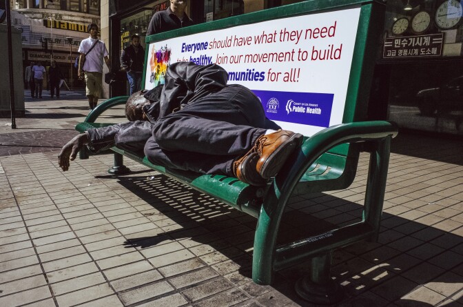 A man is lying down on a bus bench with one arm hanging over the side.
