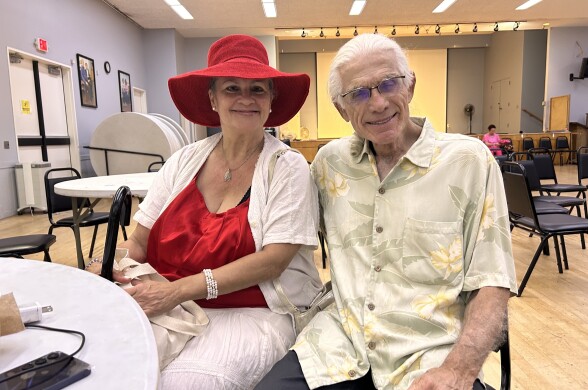 A woman, wearing a bright red hat with a long brim and a similar colored shirt, is sitting next a man with white hair, wearing a tan and white muted floral-patterned shirt.
