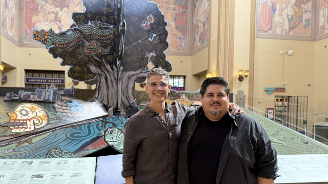 Two men pose in front of a giant pop-up-book art installation featuring a tree, a feathered serpent and a sea turtle inside the LA Central Library rotunda.