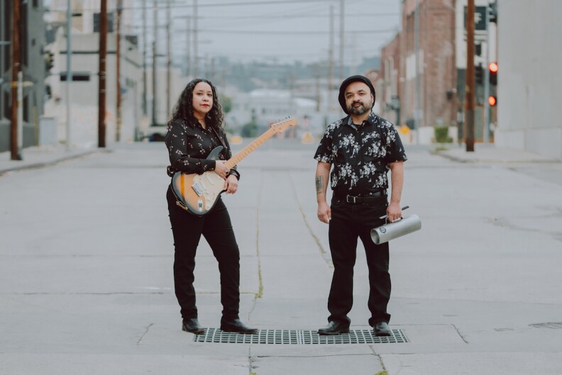 A man and woman, both wearing black, stand in the middle of an empty urban street. She holds a guitar. He holds another musical instrument.
