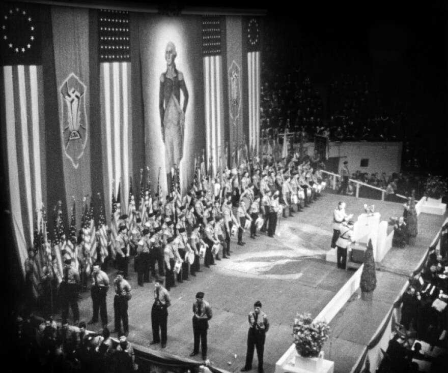 A black and white photo shows men in unforms and caps, some with instruments, others with their hands behind their backs.A speaker stands at a lectern.